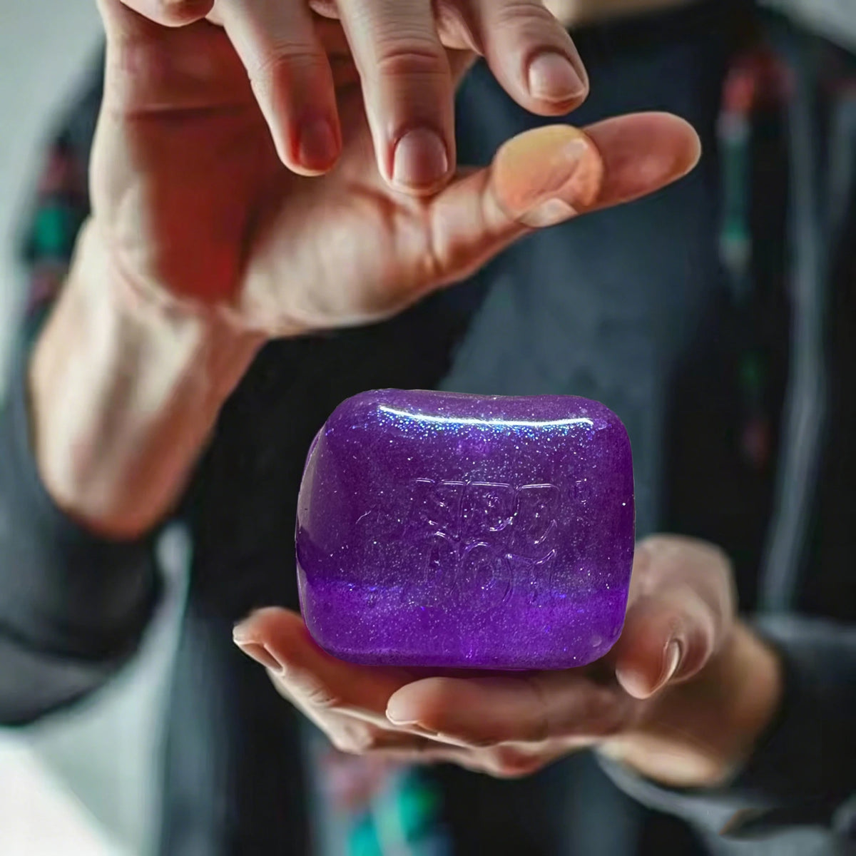 A hand holds a translucent purple glitter ice cube sensory fidget toy against a dark background.