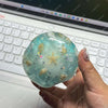 A clear, translucent blue glass stress ball filled with small shells and glitter resting on a laptop keyboard.