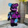 A large, black and purple knitted bear building block display model with blue accents standing next to a vase.
