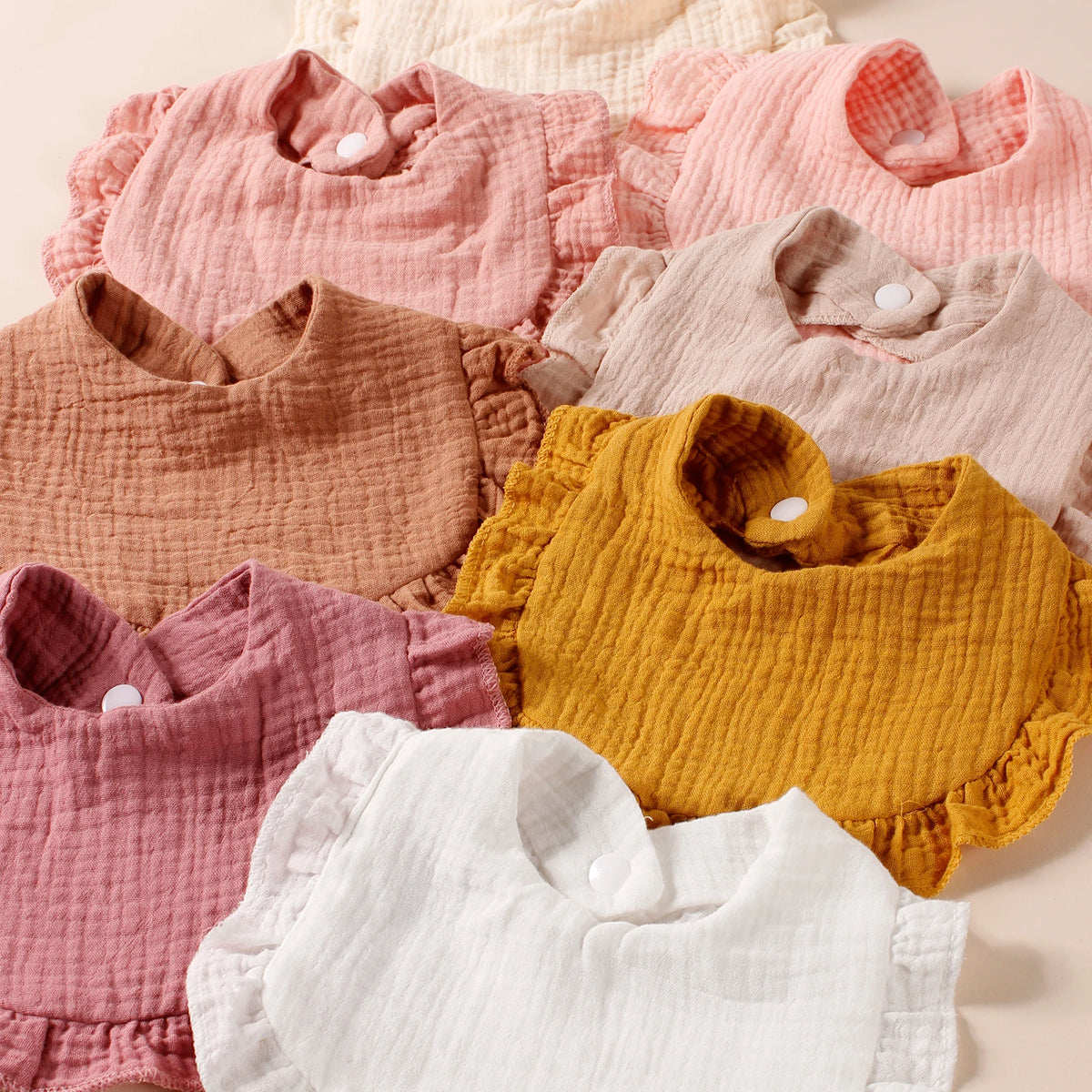 A collection of eight pink, peach, and yellow cotton ruffle baby bibs laid out on a light background.