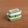A miniature doll picnic lunch box with a handle in green and white is shown on a wooden surface.