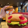 A close-up shot of a giant burger made from a plush toy, featuring an orange bun and red monster eyes.