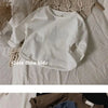 A white long sleeve cotton tee is displayed on a light fabric, with a beige knitted garment in the background.