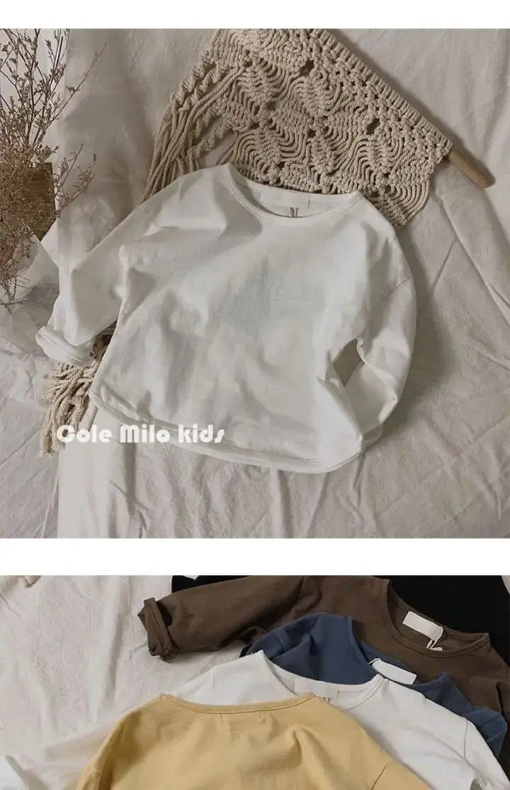 A white long sleeve cotton tee is displayed on a light fabric, with a beige knitted garment in the background.