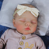 A lifelike 19-inch reborn baby doll with a soft cloth body wearing a light pink outfit and a white bow headband.