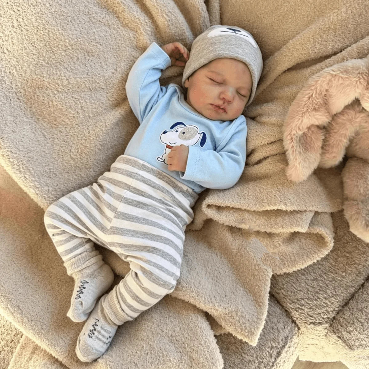 A newborn vinyl doll wearing a light blue long-sleeved top and striped pants, resting on a fluffy beige blanket.