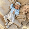 A newborn vinyl doll wearing a light blue long-sleeved top and striped pants, resting on a fluffy beige blanket.