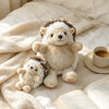 A fluffy, light brown and white hedgehog plush toy resting on a cream blanket next to an open book.