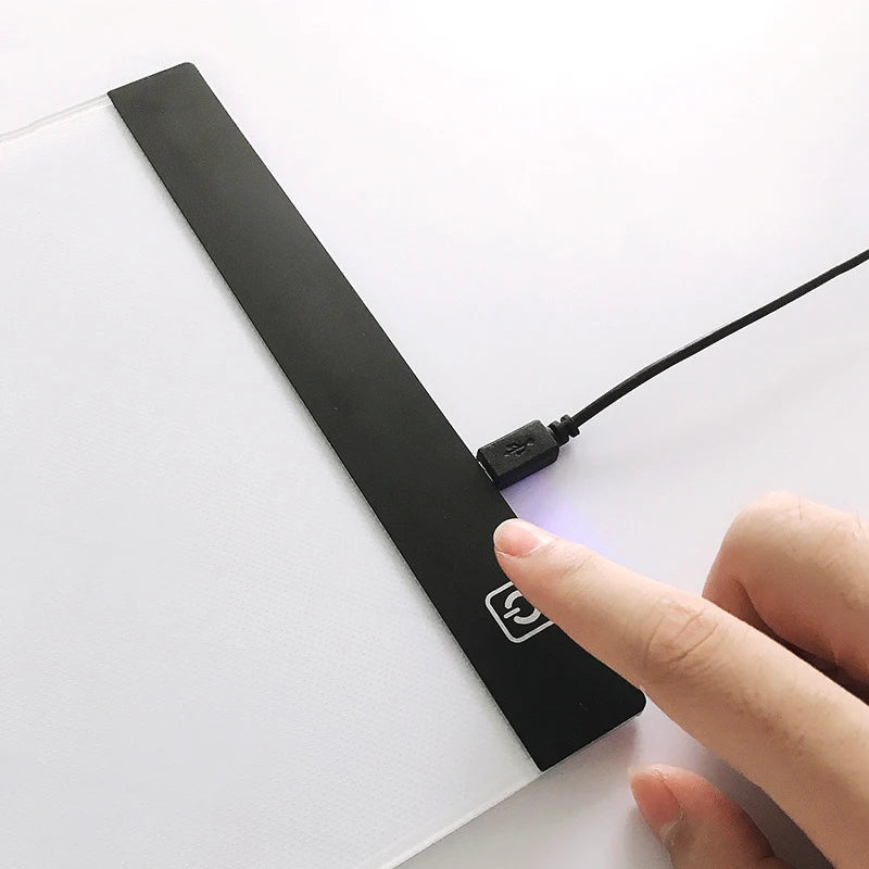 A hand is holding a black LED drawing board with a wire connected to it on a white surface.