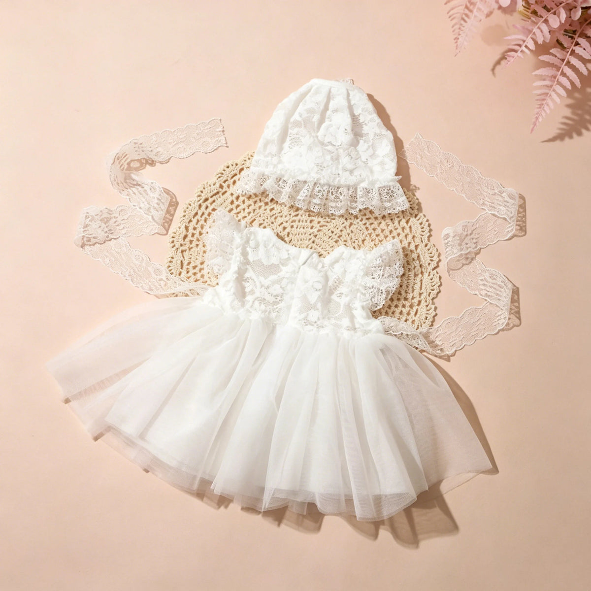 A white lace tulle dress for a newborn baby girl with a matching bonnet, displayed on a light pink background.