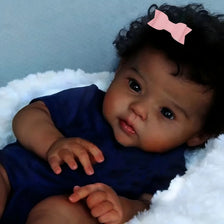 A close-up photo of a reborn baby doll with dark curly hair wearing a navy blue outfit and a pink bow, resting on white fluff