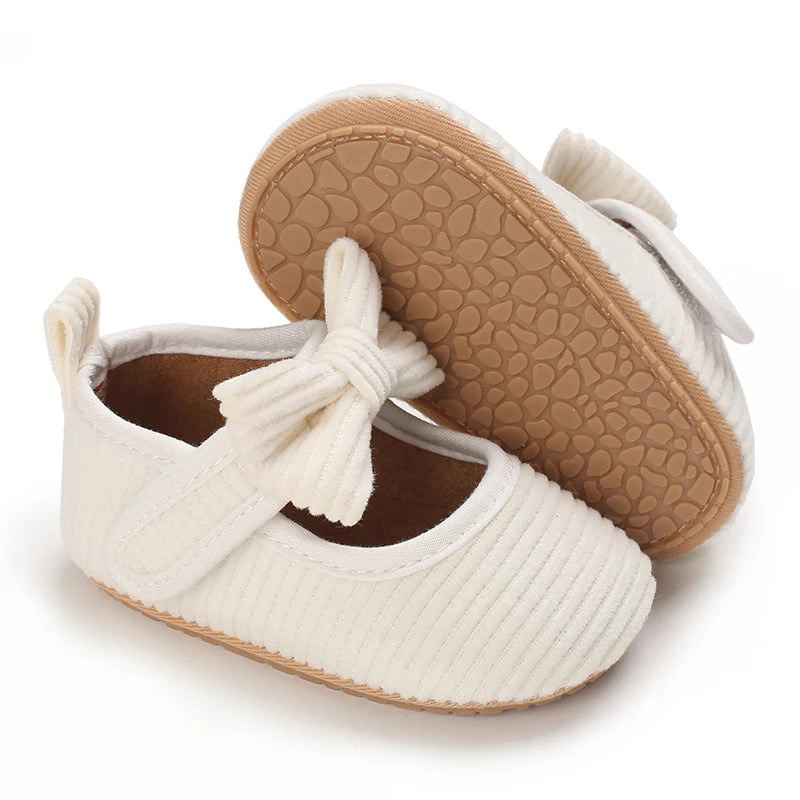 A pair of white lace-trimmed baby girl moccasin shoes with bows and tan textured soles.