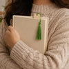 A person wearing a cream knitted sweater holds up blank wooden bookmarks with green tassels.