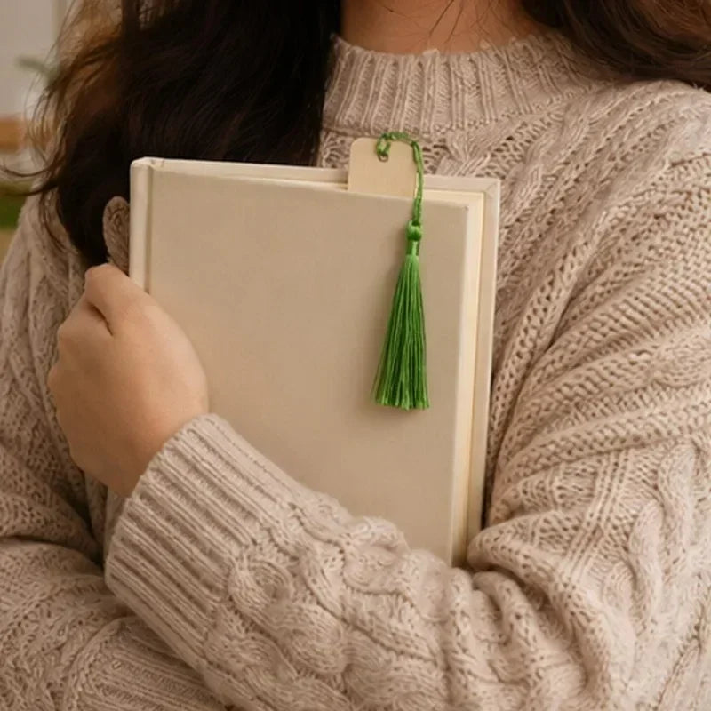 A person wearing a cream knitted sweater holds up blank wooden bookmarks with green tassels.