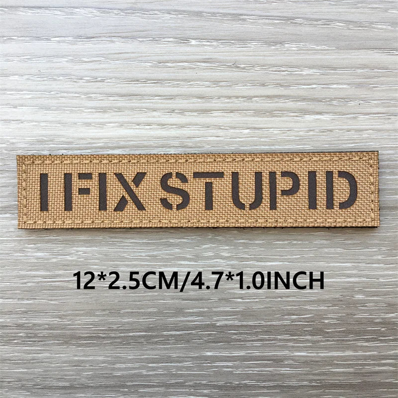 Un patch rectangulaire marron tissé avec le texte IFIXSTUPID, montré sur un fond en bois clair.