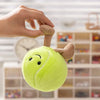A hand is holding a bright lime green plush sports ball with attached feet against a blurred background of wooden shelves.