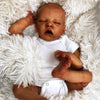 A realistic newborn twin baby doll with dark skin lying on a white fluffy blanket.