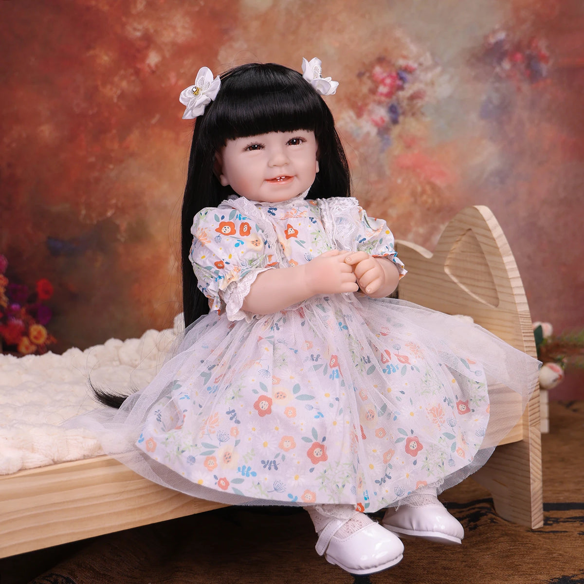 A lifelike reborn baby doll with a soft cloth body dressed in a floral white dress sitting on a wooden surface.