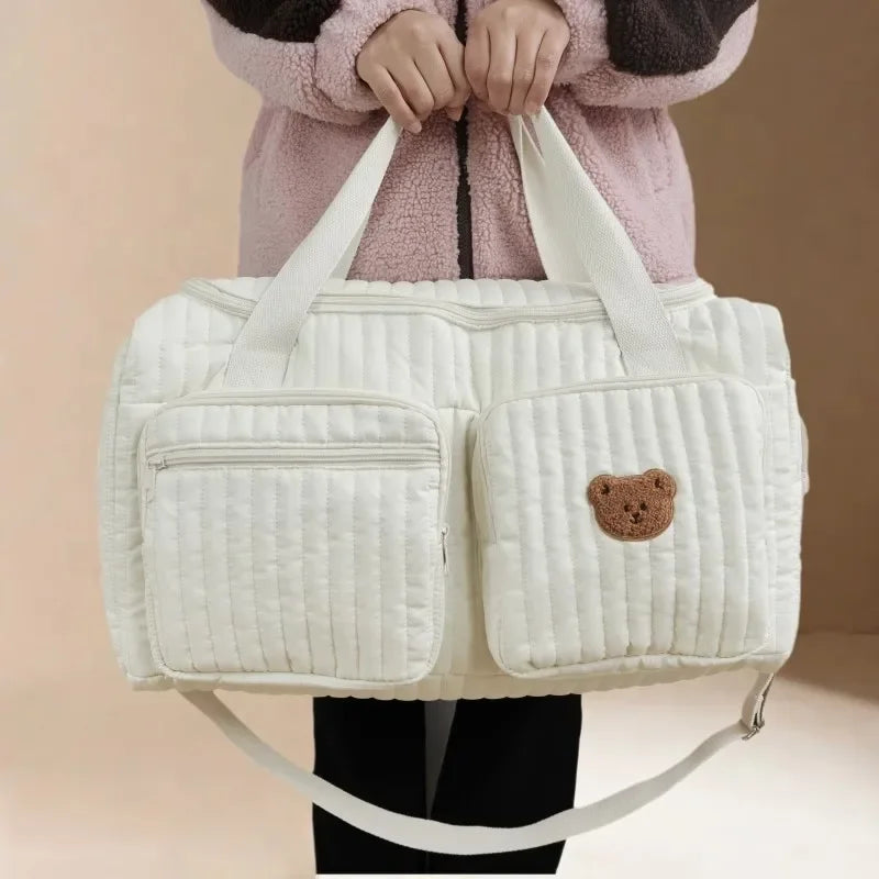 A white quilted diaper bag with brown bear patch detail is held by a person wearing a pink sweater.