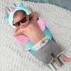 A newborn baby is lying on a light gray blanket wearing a pink and blue head protector backpack and sunglasses.