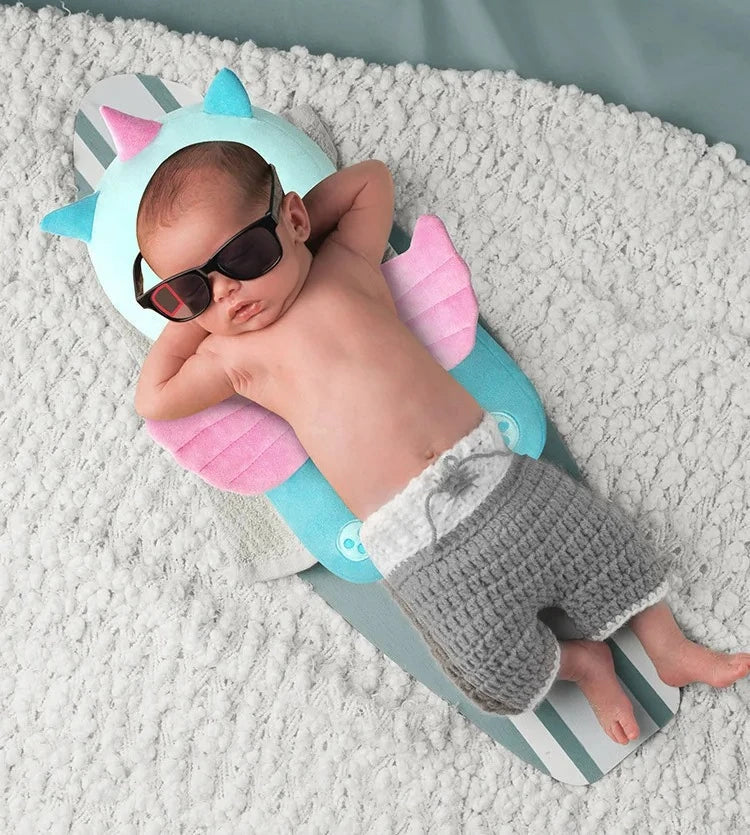 A newborn baby is lying on a light gray blanket wearing a pink and blue head protector backpack and sunglasses.