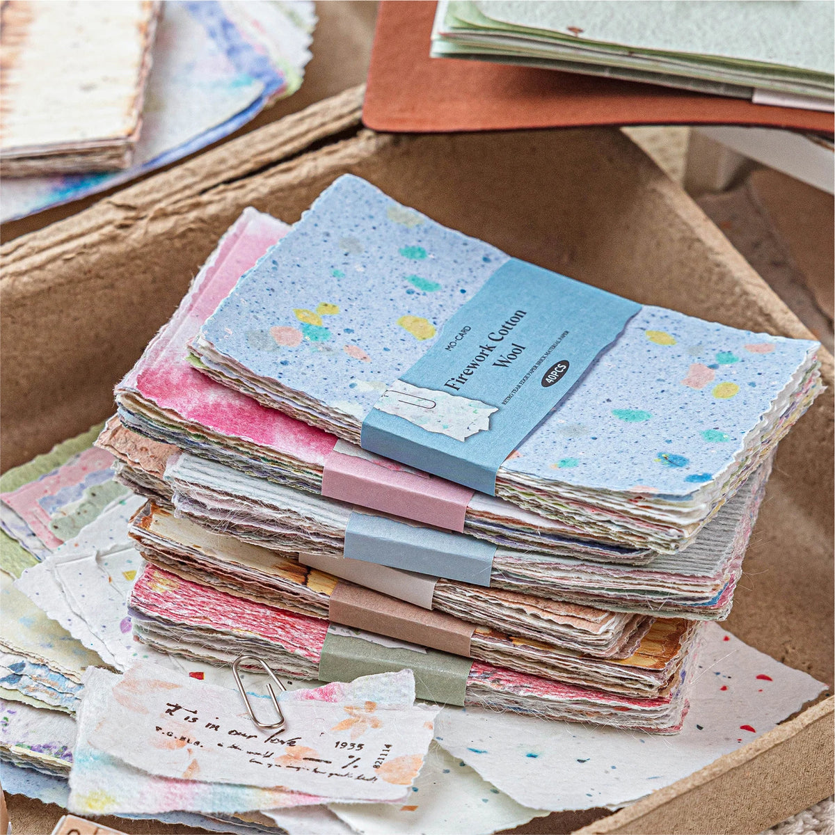 A stack of vintage scrapbook paper sheets in various colors and textures is displayed in a cardboard box.