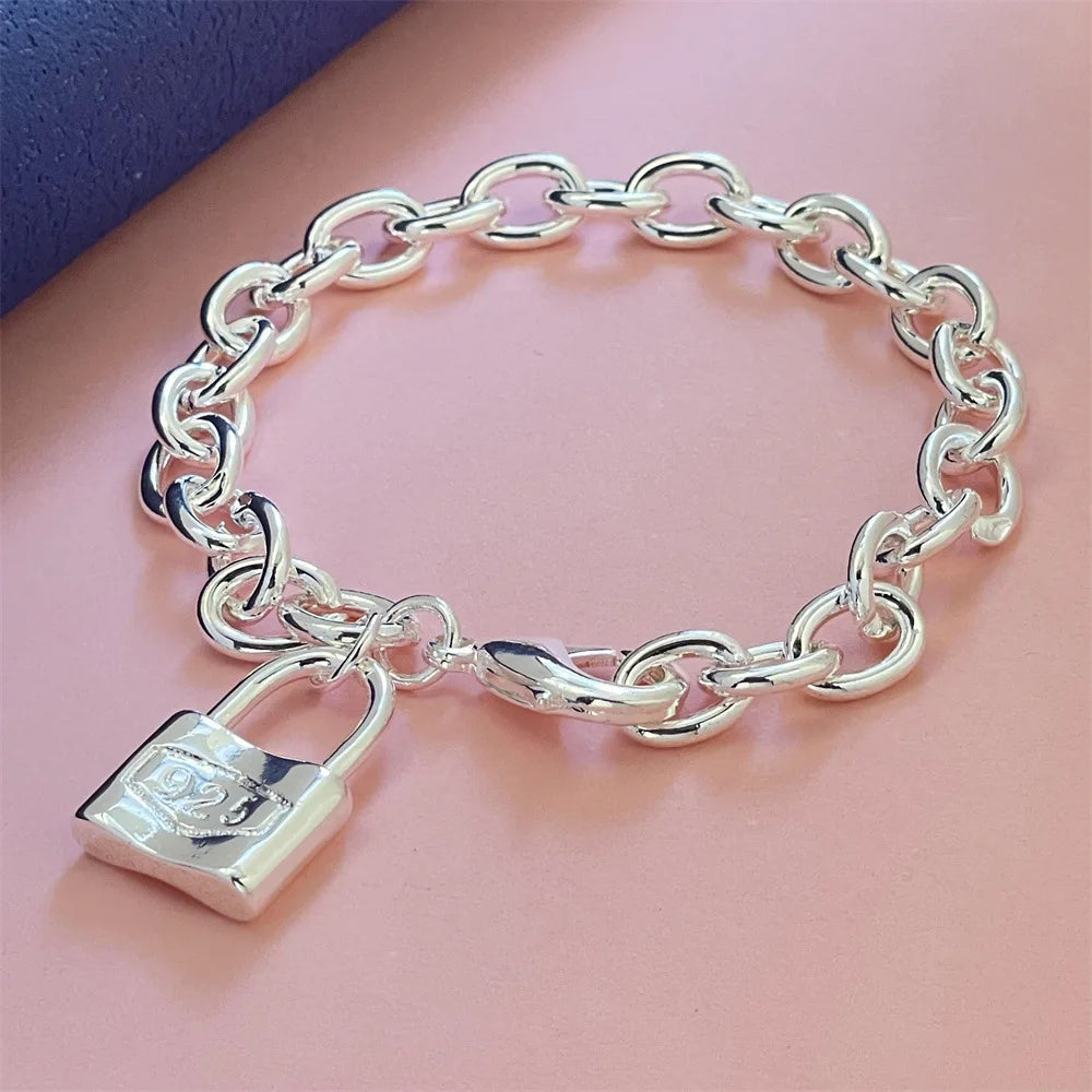 A silver snake chain bracelet with a padlock charm displayed on a pink surface.