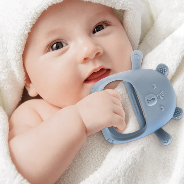 A baby is holding a silicone teether glove with a cartoon bear design while wrapped in a white towel.