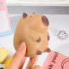 A hand holds a beige capybara squeeze stress relief toy against a pink background.
