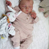 A newborn reborn baby doll with a soft cloth body wearing pink clothing and a bow, resting on white fluffy fabric.