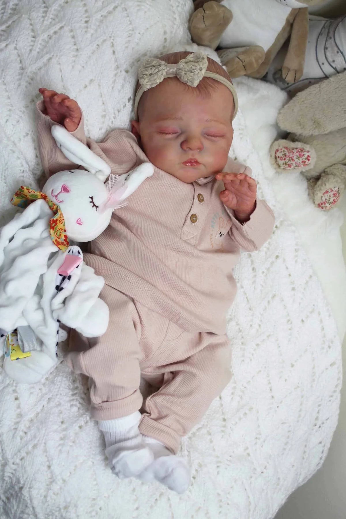 A newborn reborn baby doll with a soft cloth body wearing pink clothing and a bow, resting on white fluffy fabric.
