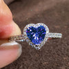 A close-up of a heart-shaped ring featuring a large, deep blue gemstone surrounded by small clear stones.