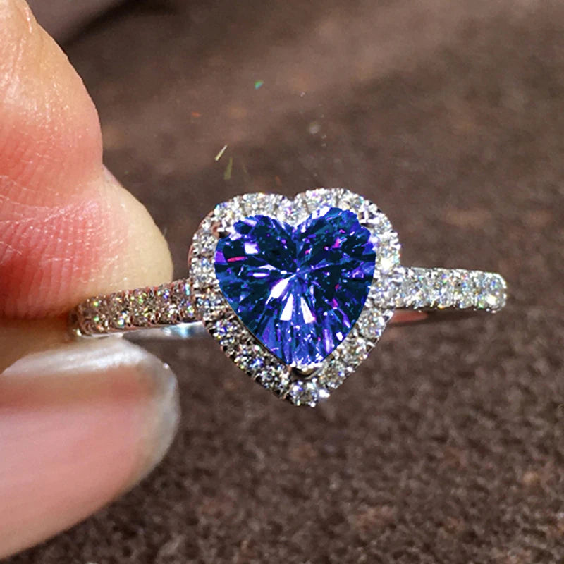 A close-up of a heart-shaped ring featuring a large, deep blue gemstone surrounded by small clear stones.