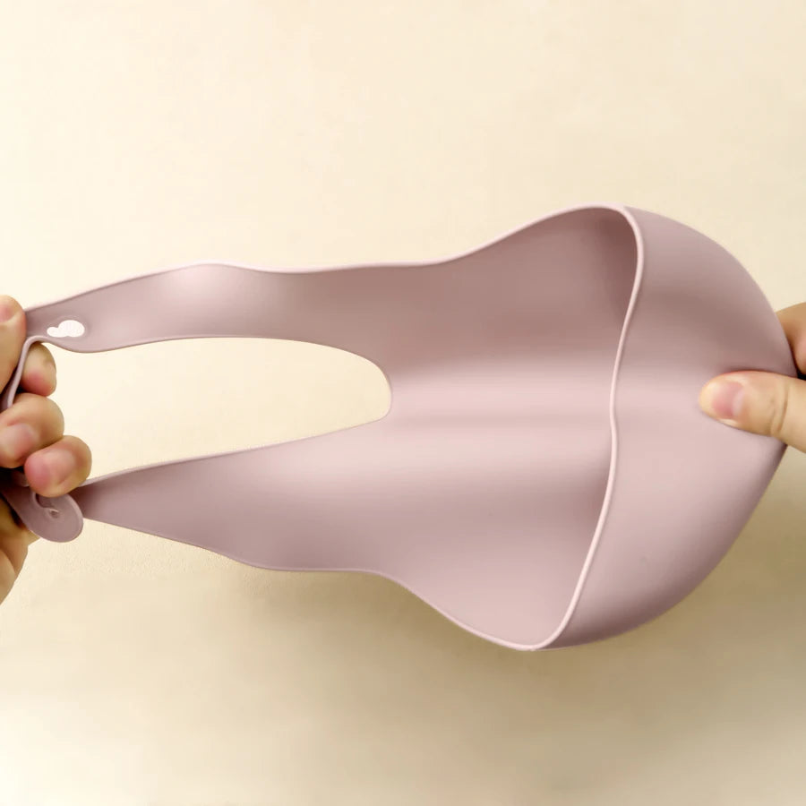 A hand holds a light pink waterproof silicone baby bib with an adjustable neck, showing the bib's shape and texture.