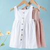 White linen jumper dress for toddler girls hanging on a wooden hanger against a light blue background