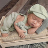 A newborn baby sleeping wearing a light green knit romper and matching newsboy cap, resting on a neutral fabric.