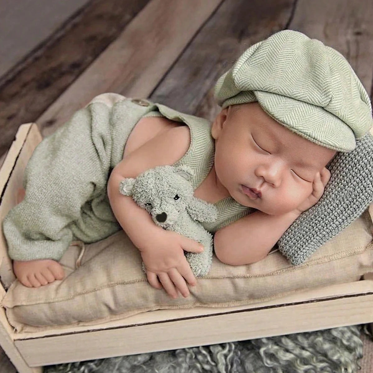 A newborn baby sleeping wearing a light green knit romper and matching newsboy cap, resting on a neutral fabric.