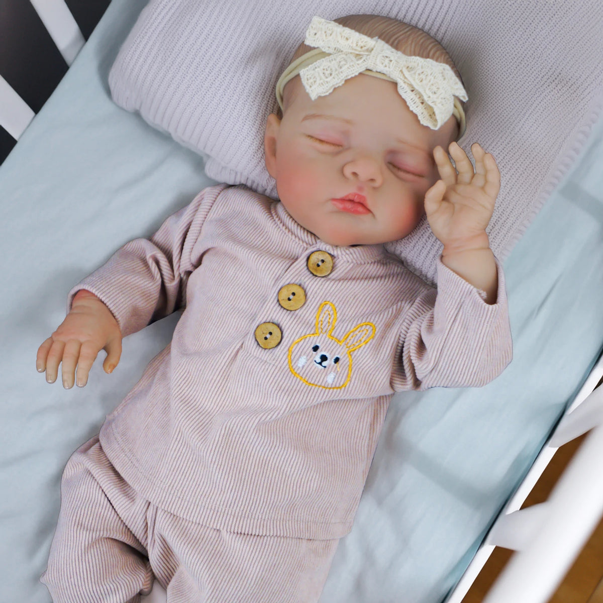 A newborn reborn baby doll with soft cloth body wearing a pink outfit and a white bow, sleeping on a light blue surface.