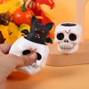 A hand holds a black cat and white skull squeeze stress ball toy against an orange background.