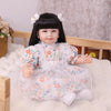 A baby doll with black hair and a floral dress is sitting on white fluffy material.