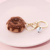 A brown crochet fruit keychain pendant with a gold-colored chain and silver bead resting on a pink surface.