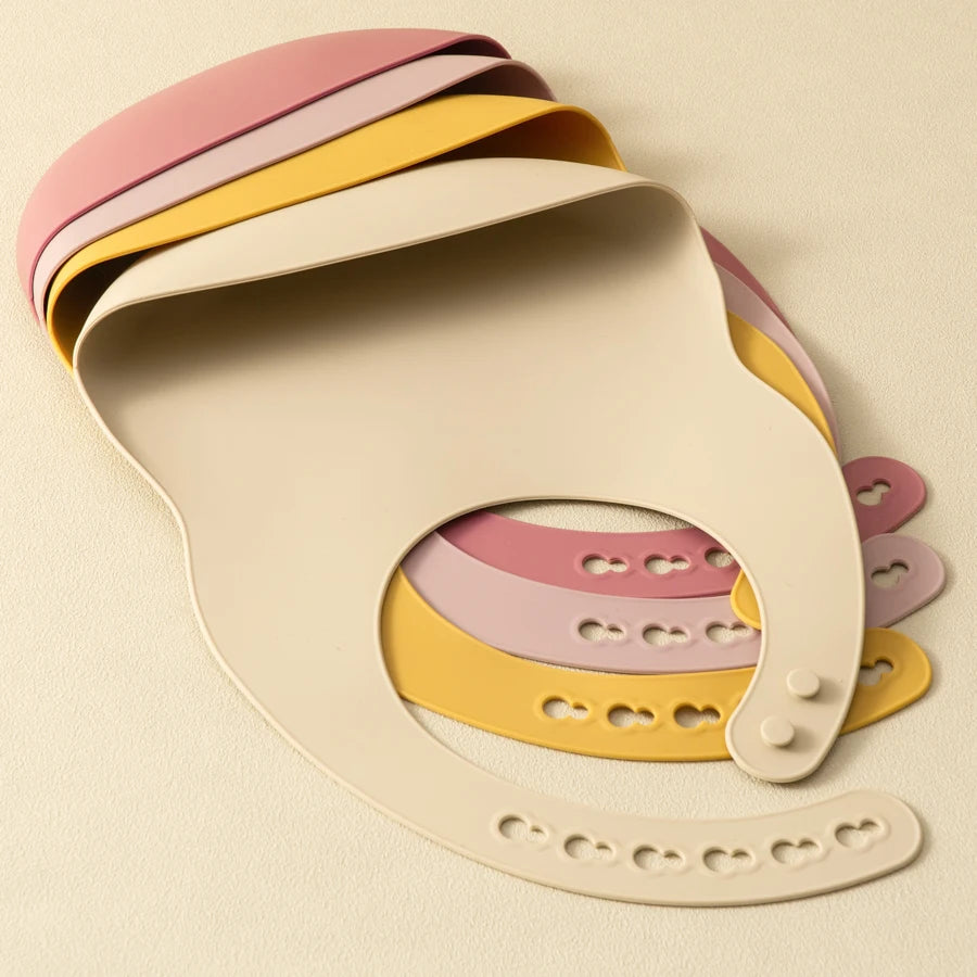 A stack of five pastel silicone baby bibs with adjustable necks, showing various shades of pink, yellow, and beige.