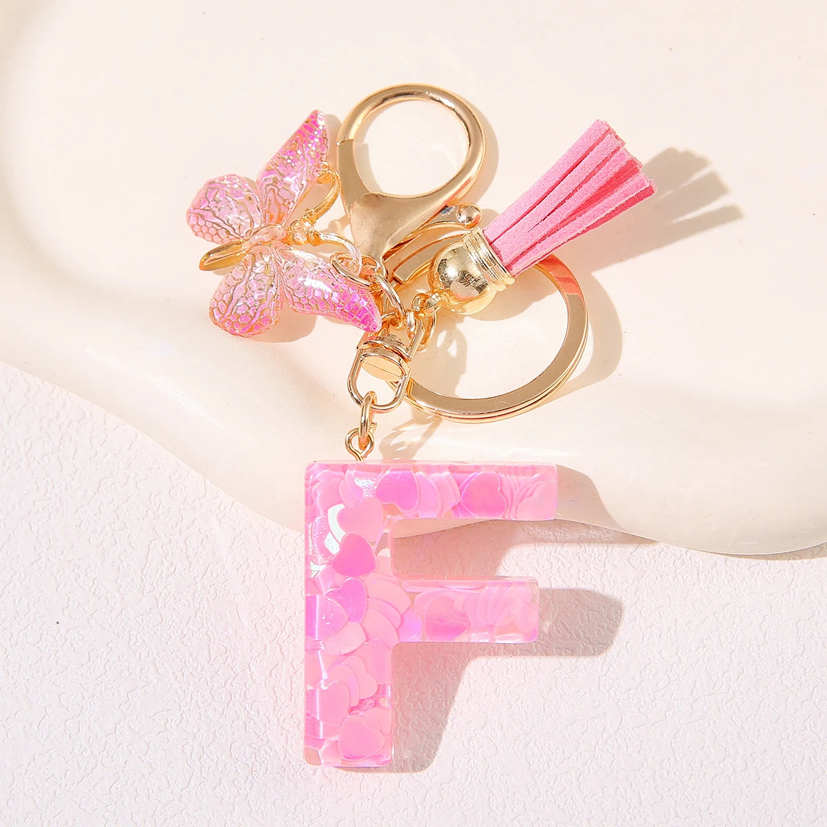 Gold initial heart keychain featuring a pink butterfly charm and a pink tassel on a white background.