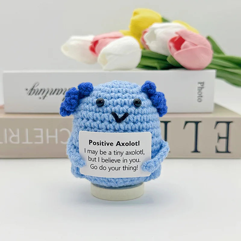 A handwoven blue crochet pencil ornament with a smiling face sits in front of a box of pencils and flowers.
