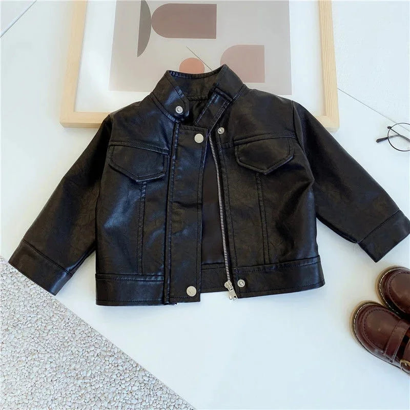 A black faux leather zip-up jacket lying on a white surface next to some brown shoes.