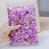A hand holds a clear plastic bag filled with multicolor resin nail charms featuring purple and pink floral designs.