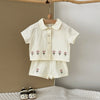 A cream baby cotton shirt and shorts set with a pink flower pattern hanging on a wooden hanger.