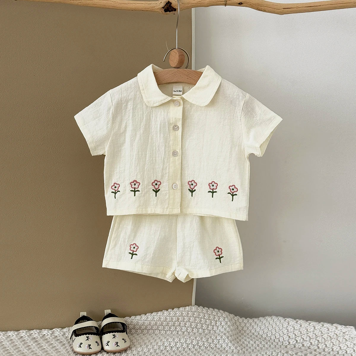 A cream baby cotton shirt and shorts set with a pink flower pattern hanging on a wooden hanger.