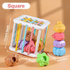 A white square toy organizer holds colorful wooden shapes and a green object for toddlers.