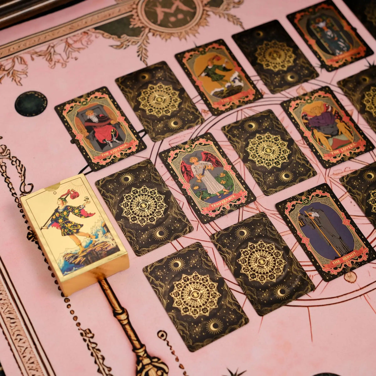 A spread of mini gold foil tarot cards laid out on pink parchment with a golden quill.